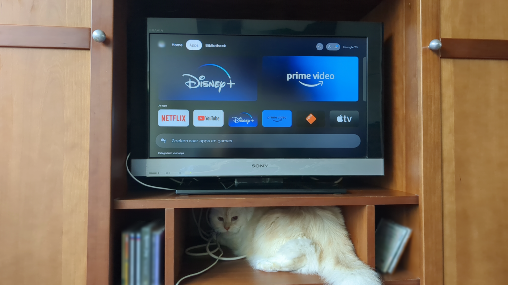 Foto van een tv waarop een Chromecast geïnstalleerd is. Het homescherm met alle apps is te zien. De tv staat in een tv-meubel waarin ook een kat is gaan zitten, die een beetje dommig richting de camera kijkt.