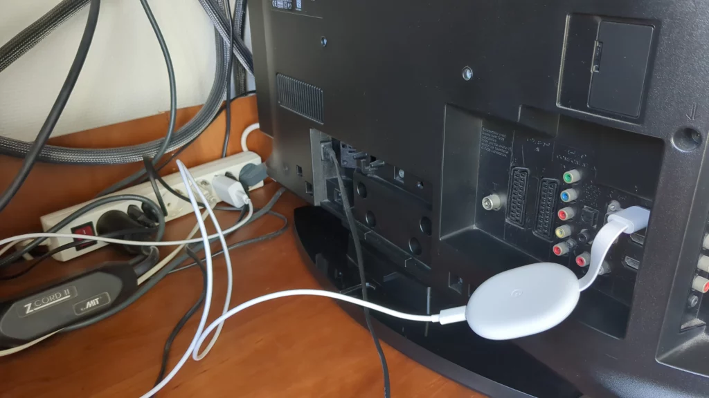 Achterkant van een tv waarin een Chromecast is geplugd in de HDMI-poort, met de bijbehorende lader aangesloten op een stekkerdoos. 